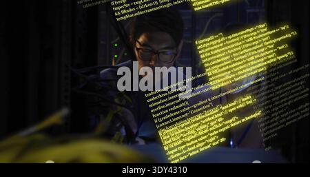 Korean IT technician wearing glasses tracing cables in dim server rack, showing yellow code overlay Stock Photo