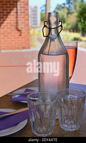 Condensation covered glass water bottle with drinking glasses on outdoor café or restaurant table in sunny garden setting Stock Photo
