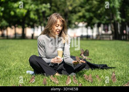 Positive businesswoman relaxing in park sitting on grass eating ice cream and feeding curious birds. Stock Photo