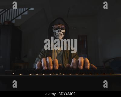 A person wearing a highly detailed skull-print face mask and a hooded jacket while typing on a glowing mechanical keyboard. Stock Photo