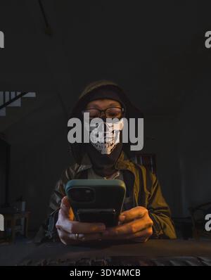 A person wearing a skull-print face mask, glasses, and a hooded jacket, actively engaged with a glowing smartphone screen. Stock Photo