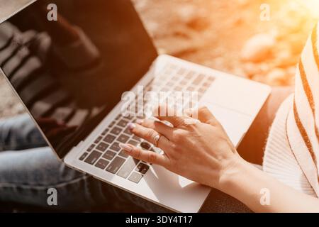 Laptop typing remote woman working on a silver laptop while sitting outside in the bright golden sunlight Stock Photo