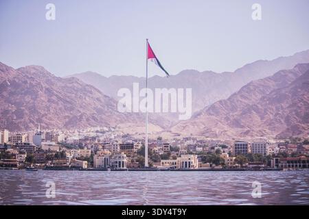 The massive Arab Revolt flag flying from the Aqaba Flagpole over the coastal city skyline and rugged desert mountains in Jordan, seen from the water. Stock Photo