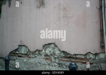 A weathered, pink-painted wall with peeling plaster exposing old brickwork, featuring a small, colorful pixel-art mosaic cat tile. Stock Photo