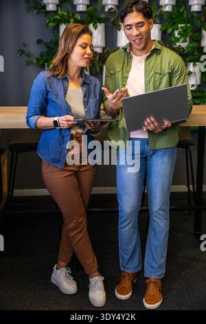 Diverse coworkers discussing project using laptop and tablet in office lounge near plant wall Stock Photo
