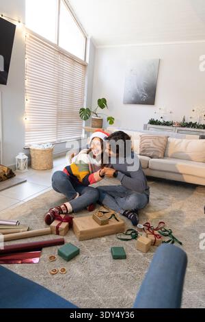 Couple sitting on rug at home wrapping gifts with rolls of paper, ribbons, wearing Santa hat Stock Photo