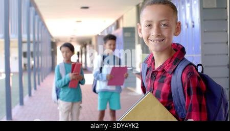 Smiling boy in red plaid shirt holding yellow folder walking school hall, with lockers, copy space Stock Photo