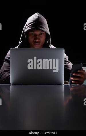 Hacker, man and laptop with phone at table for phishing, install ransomware or stealing data. Space, tech or black person with cybercrime in dark room Stock Photo