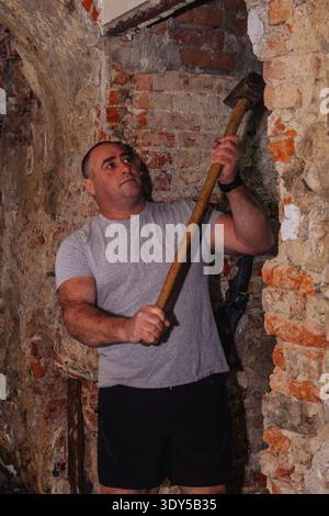 A muscular construction worker in shorts is actively breaking a brick wall with a sledgehammer on a construction site. He's focused and exerting signi Stock Photo