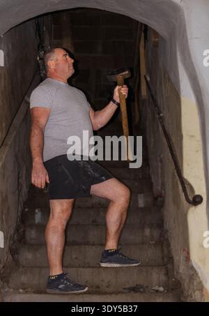 A construction worker in shorts and a gray t-shirt is descending a set of stone stairs in a building under construction. He is holding a hammer in his Stock Photo