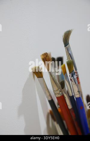 Various paintbrushes are neatly placed in a holder, showcasing different sizes and shapes, with their shadows cast on a white wall in soft natural lig Stock Photo