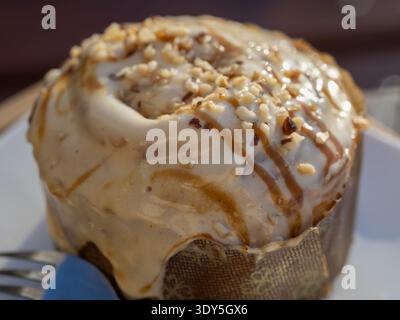 indulgent caramel glazed cinnamon roll topped with crushed hazelnuts, close-up macro of gooey icing texture on a paper baking cup, delicious sweet pas Stock Photo