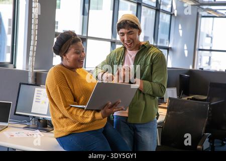 Diverse coworkers standing at workstation, leaning in and celebrating over silver laptop and charts Stock Photo