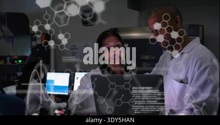 Explaining center pair wearing lab coats pointing at laptop in lab with monitors and AR overlays Stock Photo