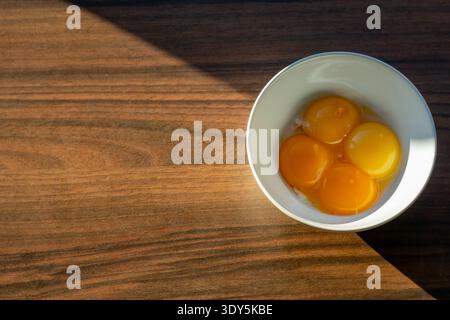 Top view of four raw egg yolks in a white bowl placed on a wooden table. Natural sunlight creates strong shadows and highlights the glossy texture. Stock Photo
