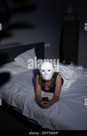 A person lies on a bed under dim light, wearing a blank mask and using a smartphone, symbolizing the importance of digital privacy and awareness of data protection against fraud. Stock Photo