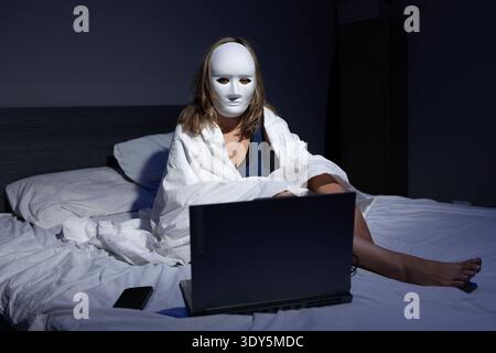 A person in a white mask is seated on a bed, wrapped in a blanket while focused on a laptop. The dark room highlights themes of privacy, data protection, and fraud prevention in the digital age. Stock Photo