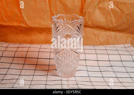 A clear glass vase featuring detailed diamond cuts and starburst designs, set against a textured orange backdrop and a white cloth with black stripes Stock Photo