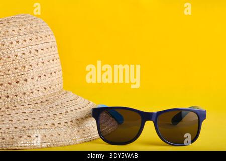 Modern blue sunglasses resting on top summer straw hat, isolated on yellow backdrop Stock Photo