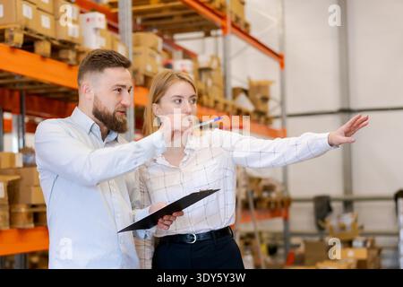 Male and female logistics managers standing together in a warehouse. Woman pointing at stock while man holds a clipboard. Concept of professional teamwork, supply chain, and inventory management. || Model released Stock Photo