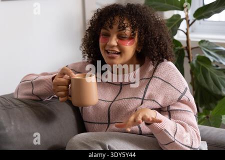 African American woman sitting on sofa at home holding mug, wearing pink sweater, red eye patches. Cozy, sunlit, plant, lounge, conversation, selfcare, wellness || Model released Stock Photo