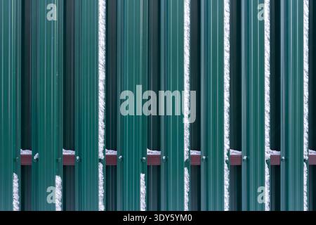 The fence is made of metal plates of green color with adhering snow. Stock Photo