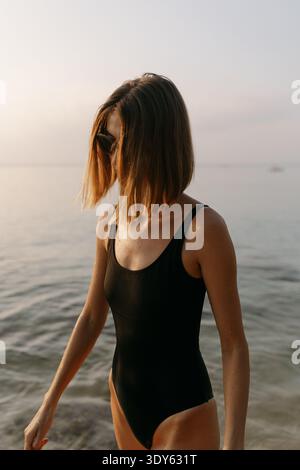 Woman in black swimsuit standing in calm sea at sunrise Stock Photo
