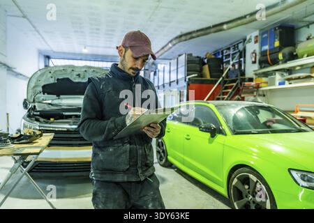 Mechanic with tattooed arms checks inspection checklist on a clipboard in a busy auto body and paint shop, repaired cars and tools visible in the back Stock Photo