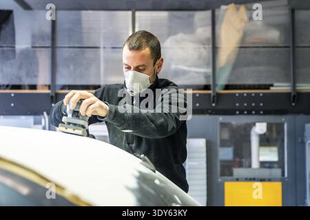 Auto body technician wearing a respirator mask using an orbital sander to prepare a car surface for painting, managing dust in a professional garage e Stock Photo