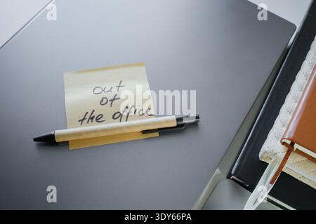Office job style flat lay. Sticky note with handwritten phrase out of the office on a laptop with pen and work notebooks . Temporarily unavailable con Stock Photo