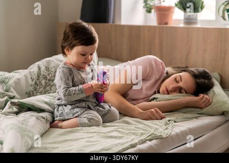 Toddler girl playing on bed while mother sleeps at home Stock Photo