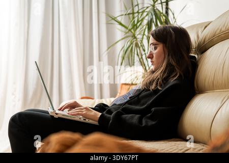 Woman student using laptop computer sitting on couch at home. Stock Photo