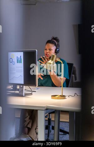 African American woman gesturing at monitor with charts and headset while sitting at office desk Stock Photo