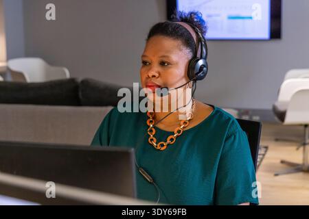 African American woman wearing headset and teal blouse sitting at desk in office using computer Stock Photo