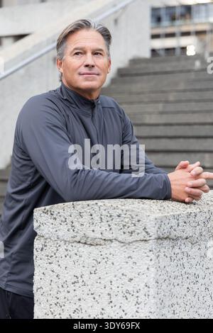 Senior man leaning on concrete post in city plaza near stairway, wearing gray jacket, looking right Stock Photo