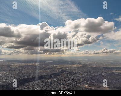 Aerial view of a sprawling city under dramatic clouds. Sunlight pierces through cloud cover, casting rays across the urban landscape. Buildings stretc Stock Photo