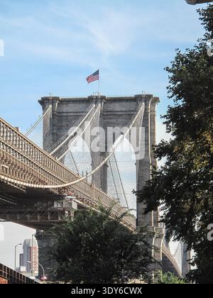 The Brooklyn Bridge spans the East River with its recognizable stone towers and steel suspension cables. An American flag flies atop one of the towers. Stock Photo