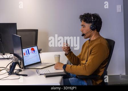 Indian man in his 20s sitting at office desk analyzing laptop charts with headset, copy space Stock Photo