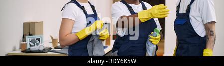 Three young adult women, one Black and two Caucasian, wearing work uniforms and yellow gloves, cleaning office space together, holding cleaning cloths and spray bottle, collaborating on tasks Stock Photo