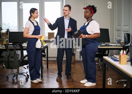 Caucasian young adult man instructing Caucasian young adult woman and Black young adult woman in cleaning uniforms, holding spray bottle and clipboard in modern office workspace Stock Photo