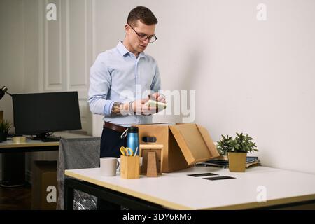 Young adult Caucasian man unpacking cardboard box on office desk, holding green object in hands, standing near workspace with stationery, mug, water bottle, and potted plant visible Stock Photo