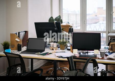 Empty office workspace showing two desks with computers, scattered documents, office supplies, potted plants, and cleaning supplies visible, suggesting recent or ongoing cleaning activity Stock Photo