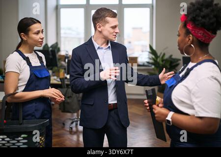 Caucasian young adult man instructing Black young adult woman and Caucasian young adult woman in cleaning uniforms holding cleaning supplies in modern office environment Stock Photo