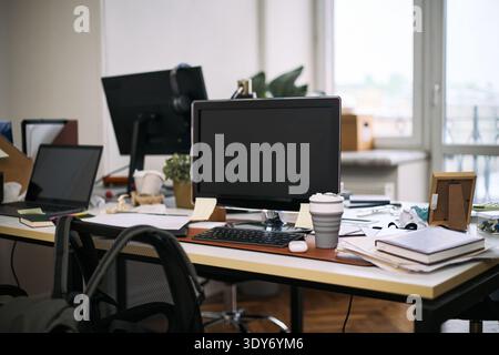 Empty office workspace showing computer monitor, keyboard, reusable coffee cup, scattered documents and supplies on desk, cleaning context implied by absence of people and organized clutter Stock Photo
