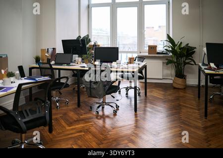 Empty modern office showing multiple workstations with computer monitors, chairs, scattered office supplies, potted plant in corner, clean hardwood floor, large windows providing natural light Stock Photo