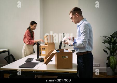 Young Caucasian man and young Asian woman cleaning office desks by packing items into cardboard boxes, both focused on organizing workspace, modern office environment visible Stock Photo