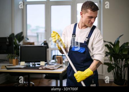 Young adult Caucasian man cleaning office floor with mop, wearing protective gloves and work overalls, standing near desk with computer and large windows in modern workspace Stock Photo