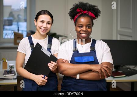 Two young adult women, one Caucasian and one Black, standing in modern office wearing cleaning uniforms, smiling at camera, one holding clipboard, demonstrating teamwork in workplace Stock Photo