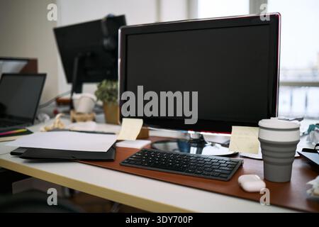 Empty office desk showing computer monitor, keyboard, wireless mouse, reusable coffee cup, folders, sticky notes and other work essentials, workspace appearing recently cleaned Stock Photo
