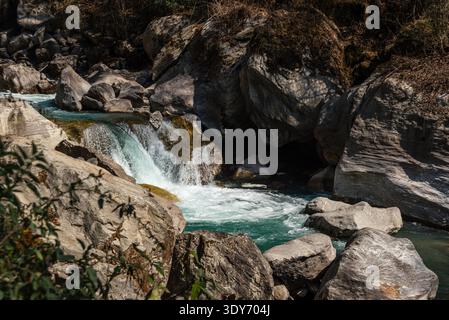 Turquoise river flows over smooth boulders in the mountains of Langtang Valley, Nepal with dry vegetation around Stock Photo
